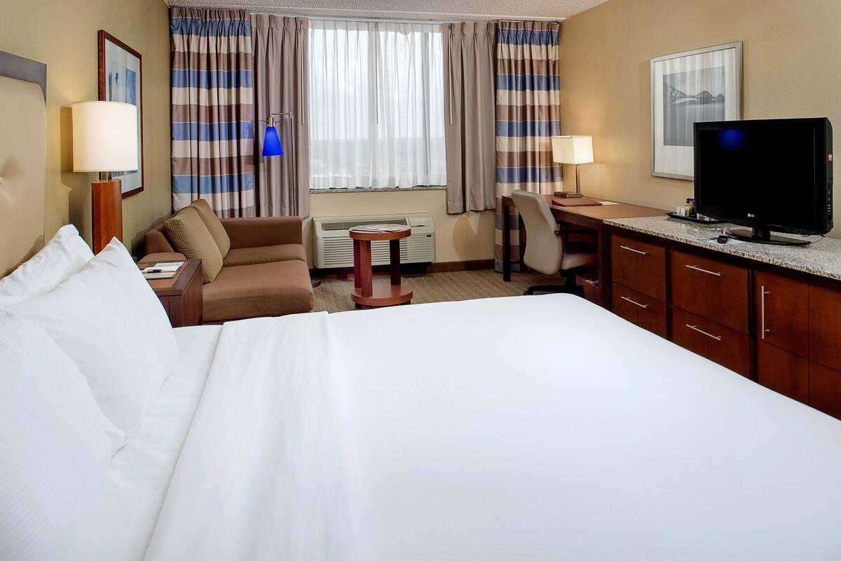 Habitación de hotel con cama, escritorio, silla, televisor y ventana con cortinas.