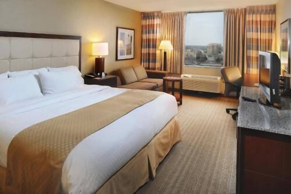 Habitación de hotel con una cama grande, un escritorio, una silla, un sofá, una lámpara y una ventana con cortinas.