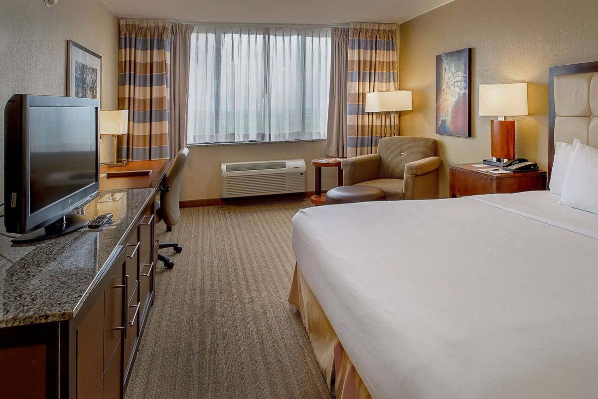 Habitación de hotel con cama, escritorio con televisor, silla, lámpara y ventana con cortinas.