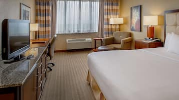 Habitación de hotel con cama, escritorio con televisor, silla, lámpara y ventana con cortinas.