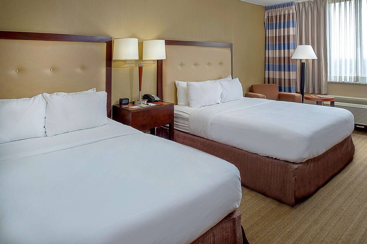 Habitación de hotel con dos camas, una mesita de noche con teléfono y lámpara, una silla y una ventana con cortinas.