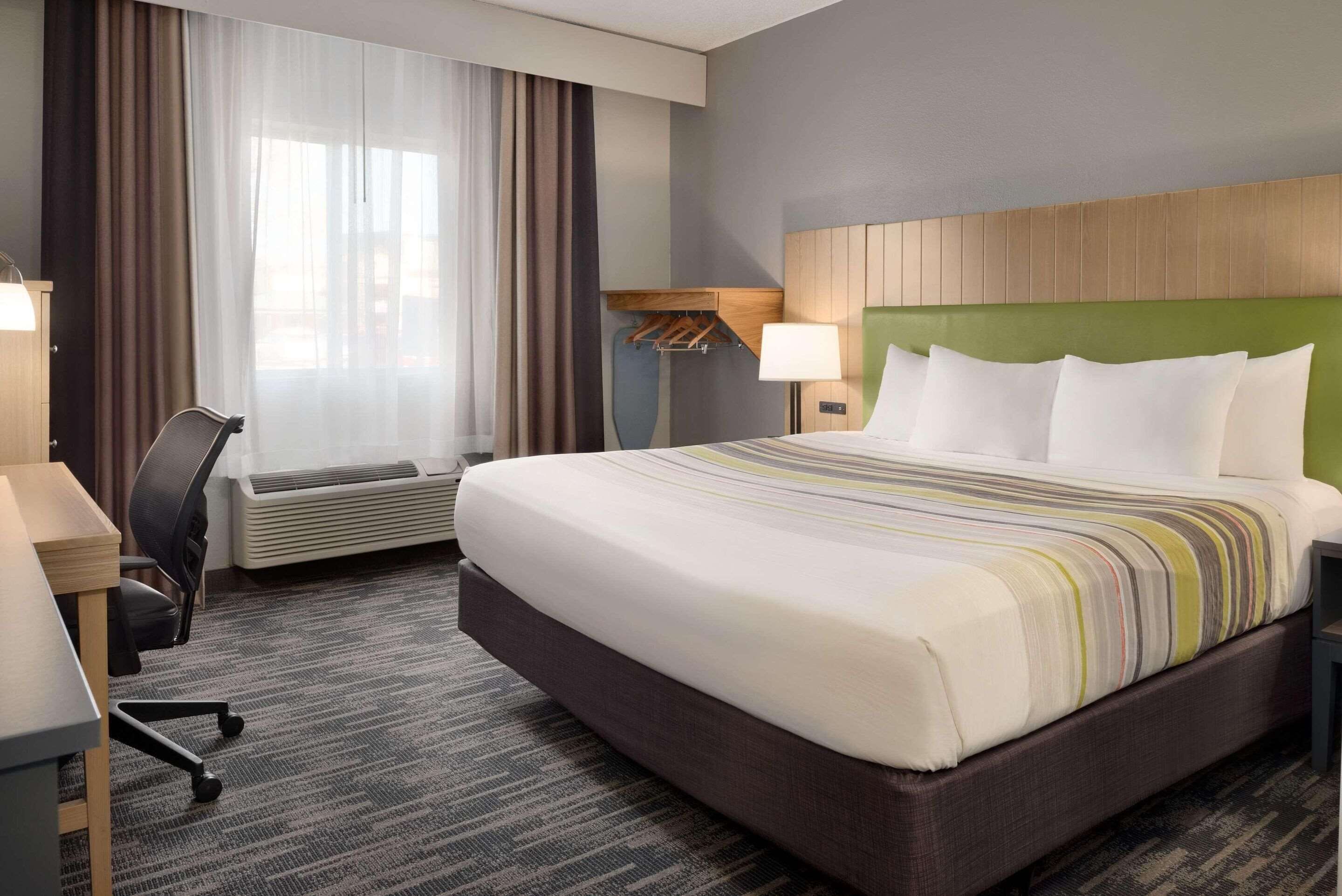 A hotel room with a large bed, a desk with a chair, a window with curtains, and a wall-mounted lamp.