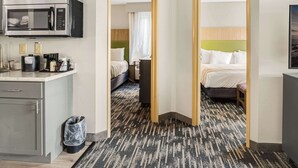A hotel room with a kitchenette, a bed with a green headboard, and a patterned carpet.
