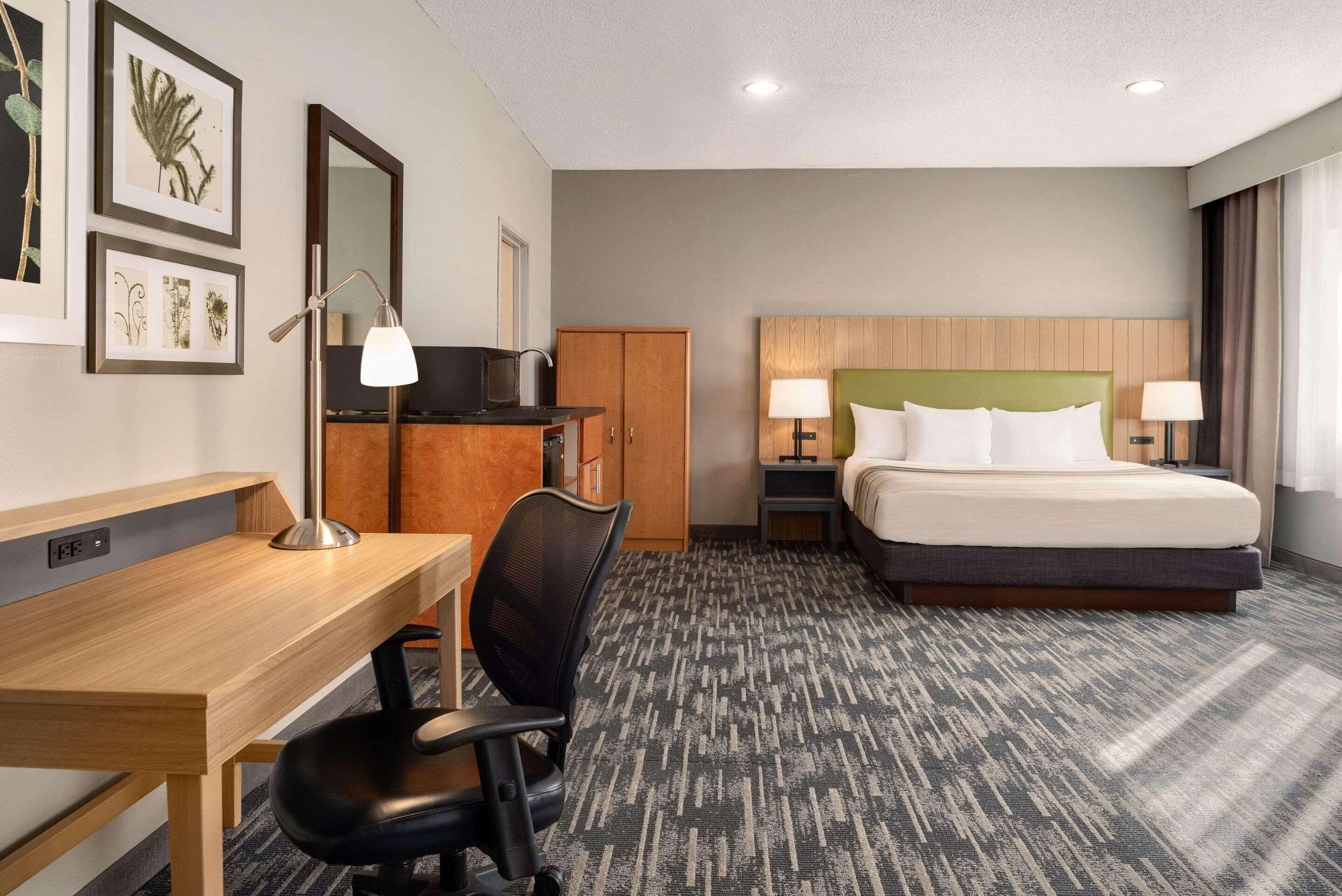 Une chambre d’hôtel avec un bureau en bois, une chaise de bureau noire, un lit avec du linge de lit blanc, deux tables de chevet avec des lampes et une armoire.