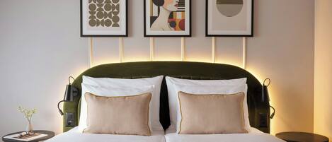 A neatly made bed with two pillows, a bedside table with a vase, and three framed artworks on the wall.