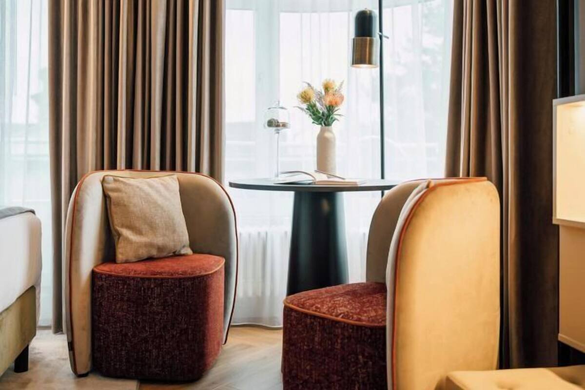 Een hotelkamer met twee fauteuils, een kleine tafel met een bloemenvaas en uitzicht op de stad door grote ramen.