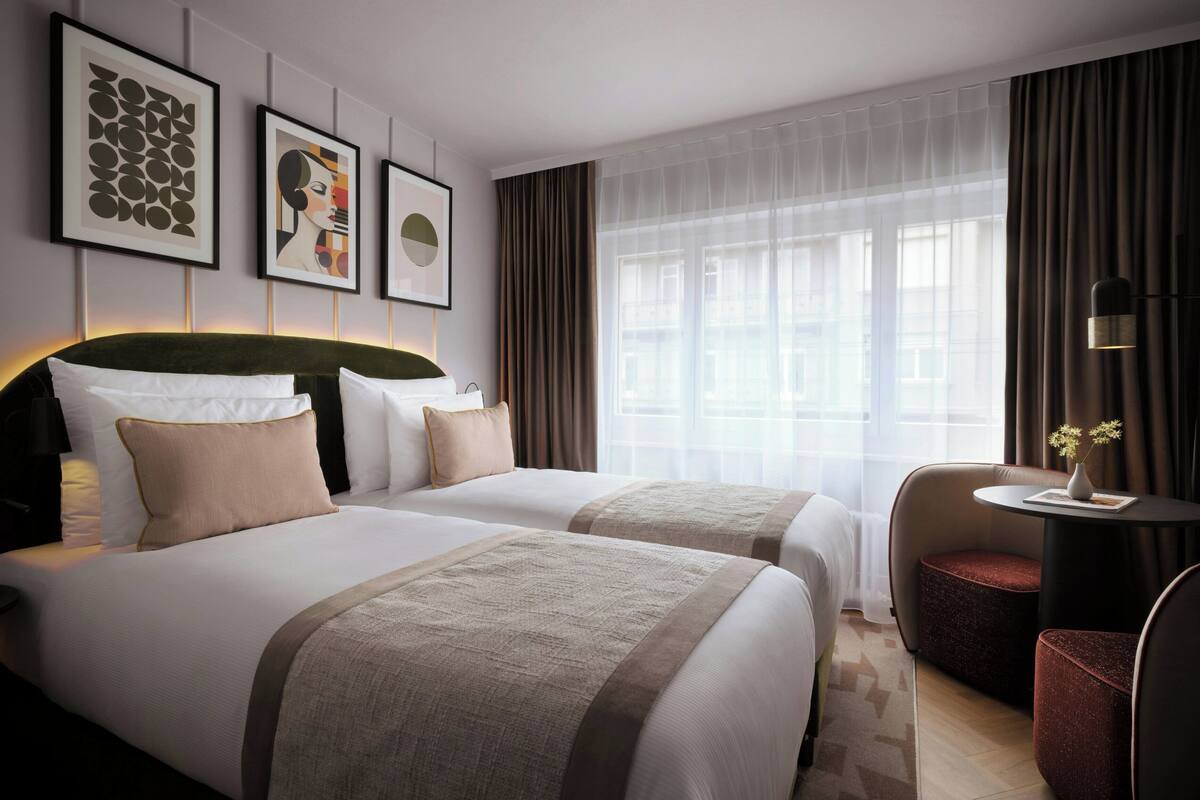 Een hotelkamer met twee bedden, een bureau met stoel, een raam met gordijnen, en kunst aan de muur.