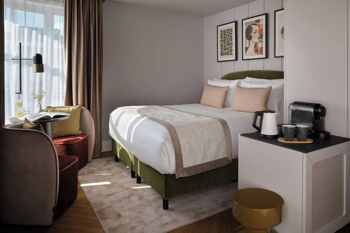 Een hotelkamer met een bed, nachtkastje, lamp, koffiezetapparaat en kunst aan de muur.