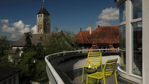 Een balkon met twee stoelen en een kleine tafel, met uitzicht op een gebouw met een klokkentoren en rode pannendaken.
