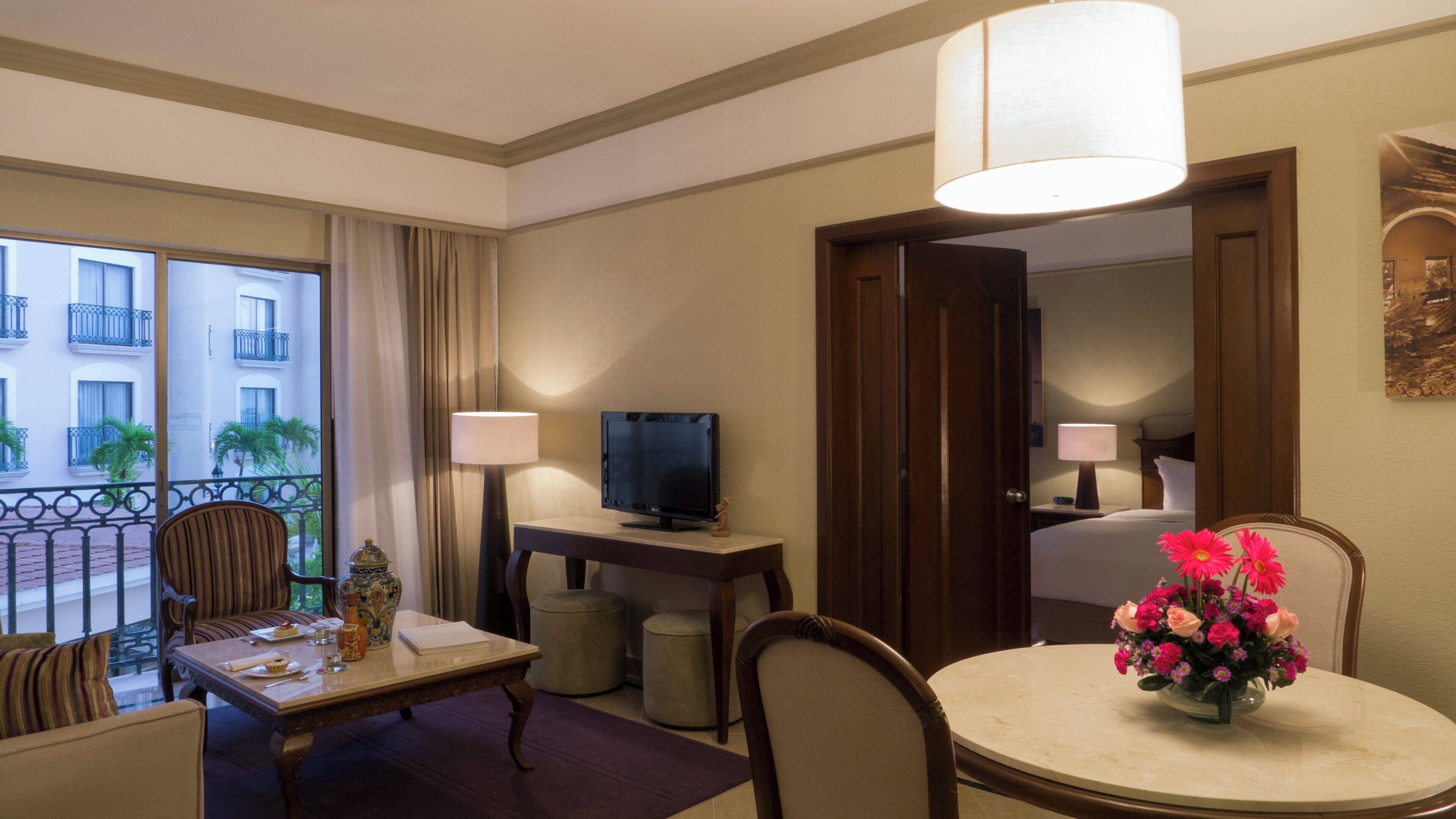 A hotel room with a balcony, a television on a desk, a dining table with chairs, and a vase of flowers.