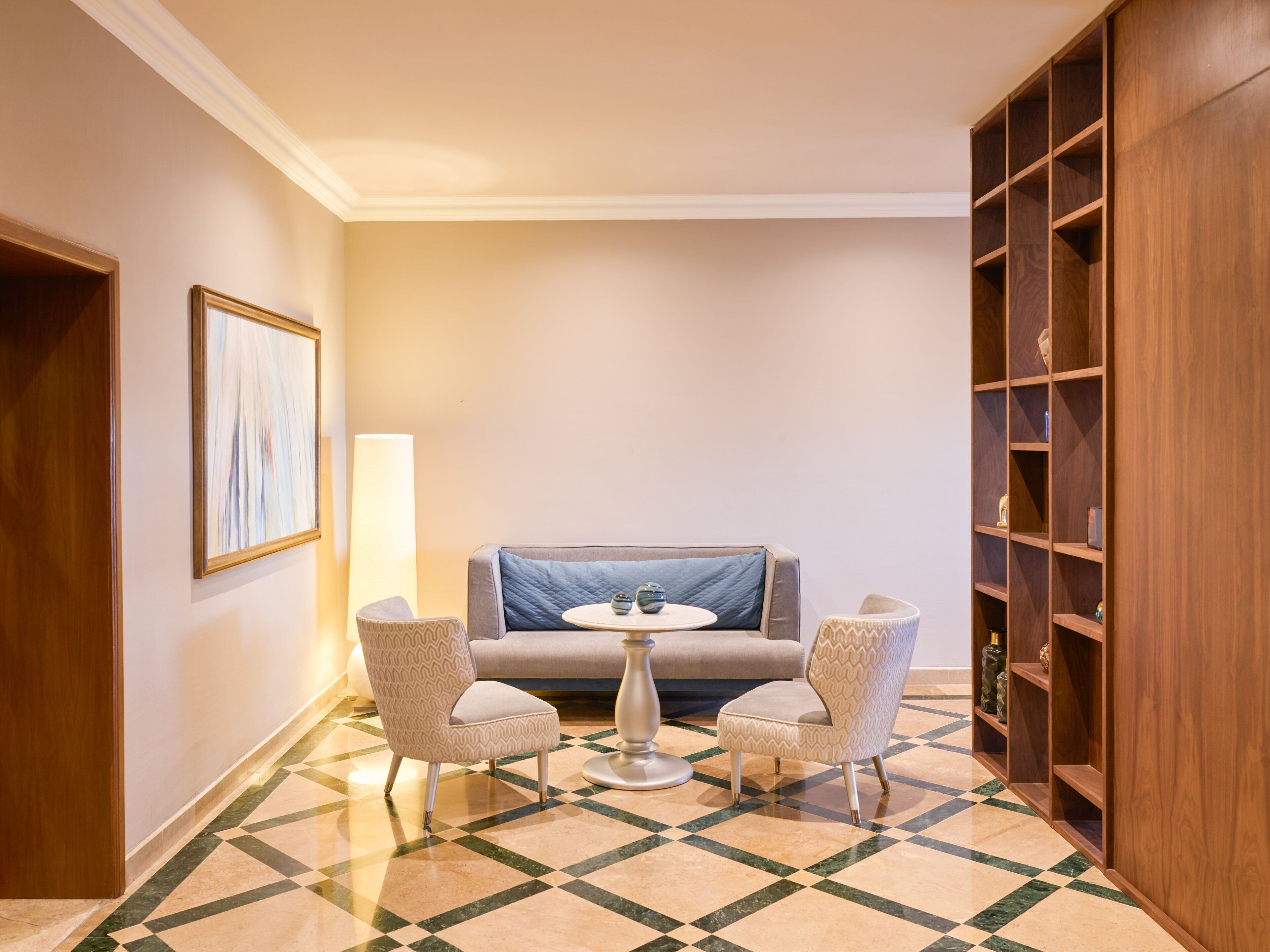 A hotel room with a checkered floor, a sofa, two patterned chairs, a small round table, and a bookshelf.