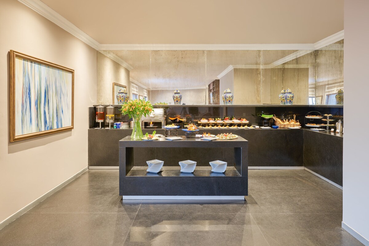 A modern hotel lobby with a reception desk, a large abstract painting, and a buffet area with various food items.