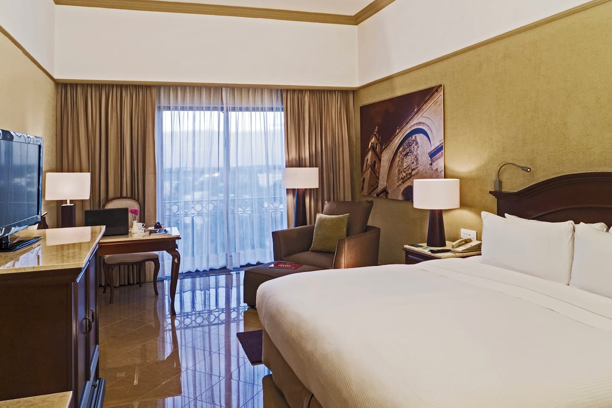 A hotel room with a large bed, a desk with a computer, a chair, a TV, and a window with curtains.