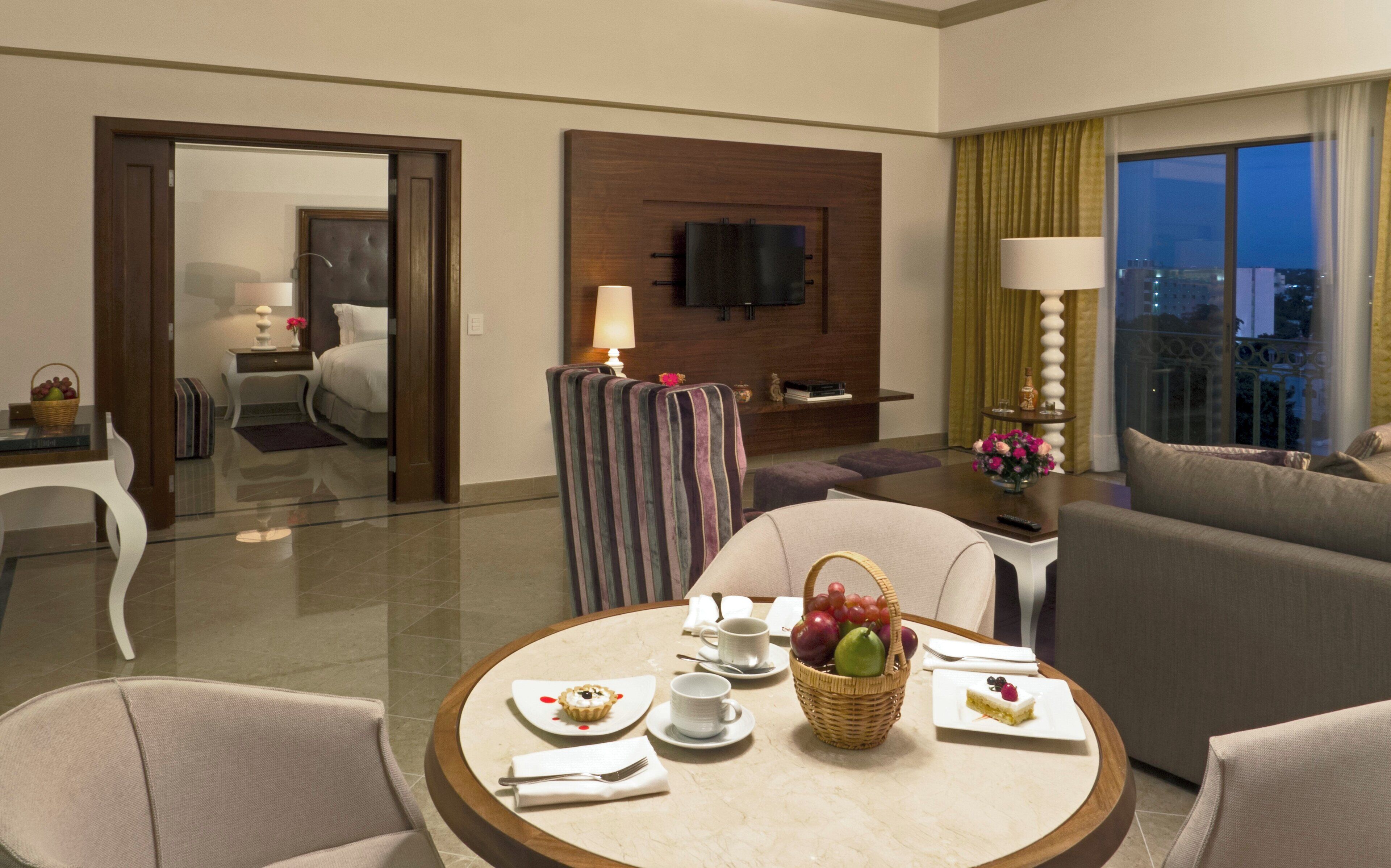 Habitación de hotel con zona de comedor, cama, televisión y vista de la ciudad de noche.