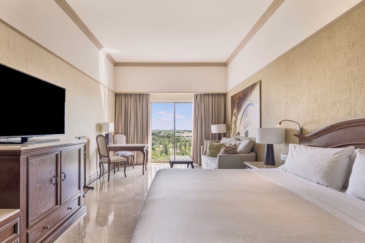A hotel room with a large bed, a television, a desk with a chair, a balcony with curtains, and a painting on the wall.