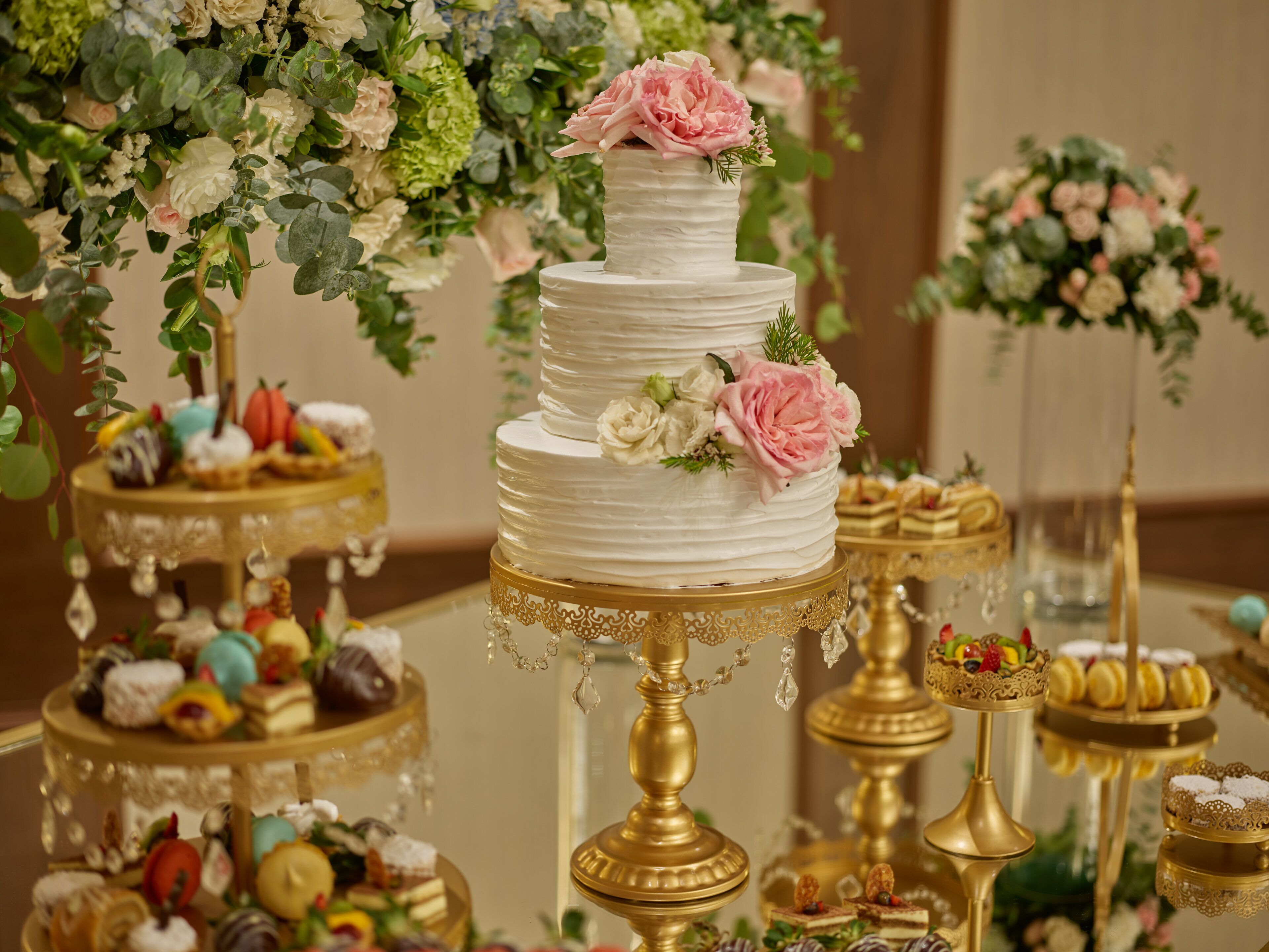 A multi-tiered wedding cake with floral decorations on a golden stand, surrounded by various dessert displays.
