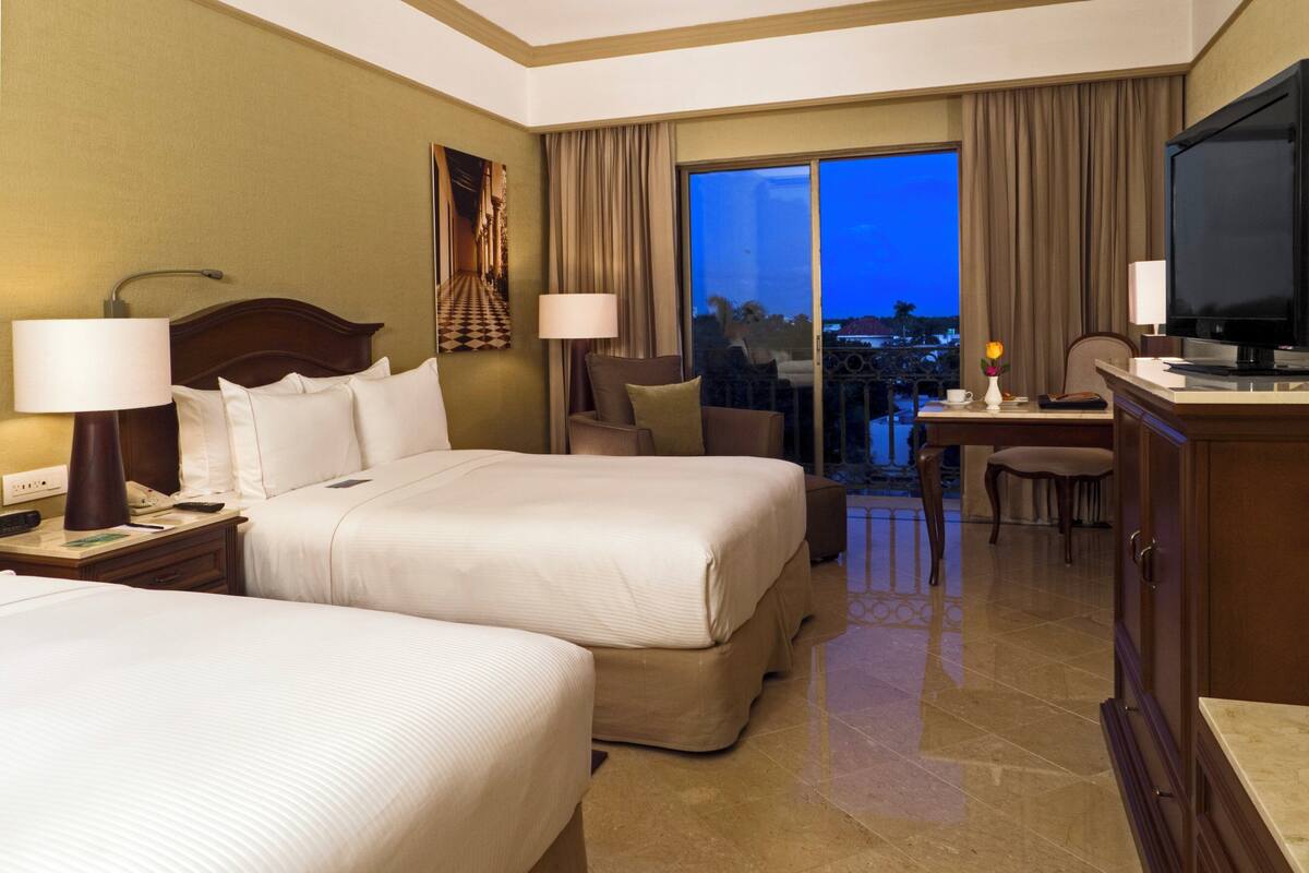 A hotel room with two beds, a desk, a television, and a balcony with a view.