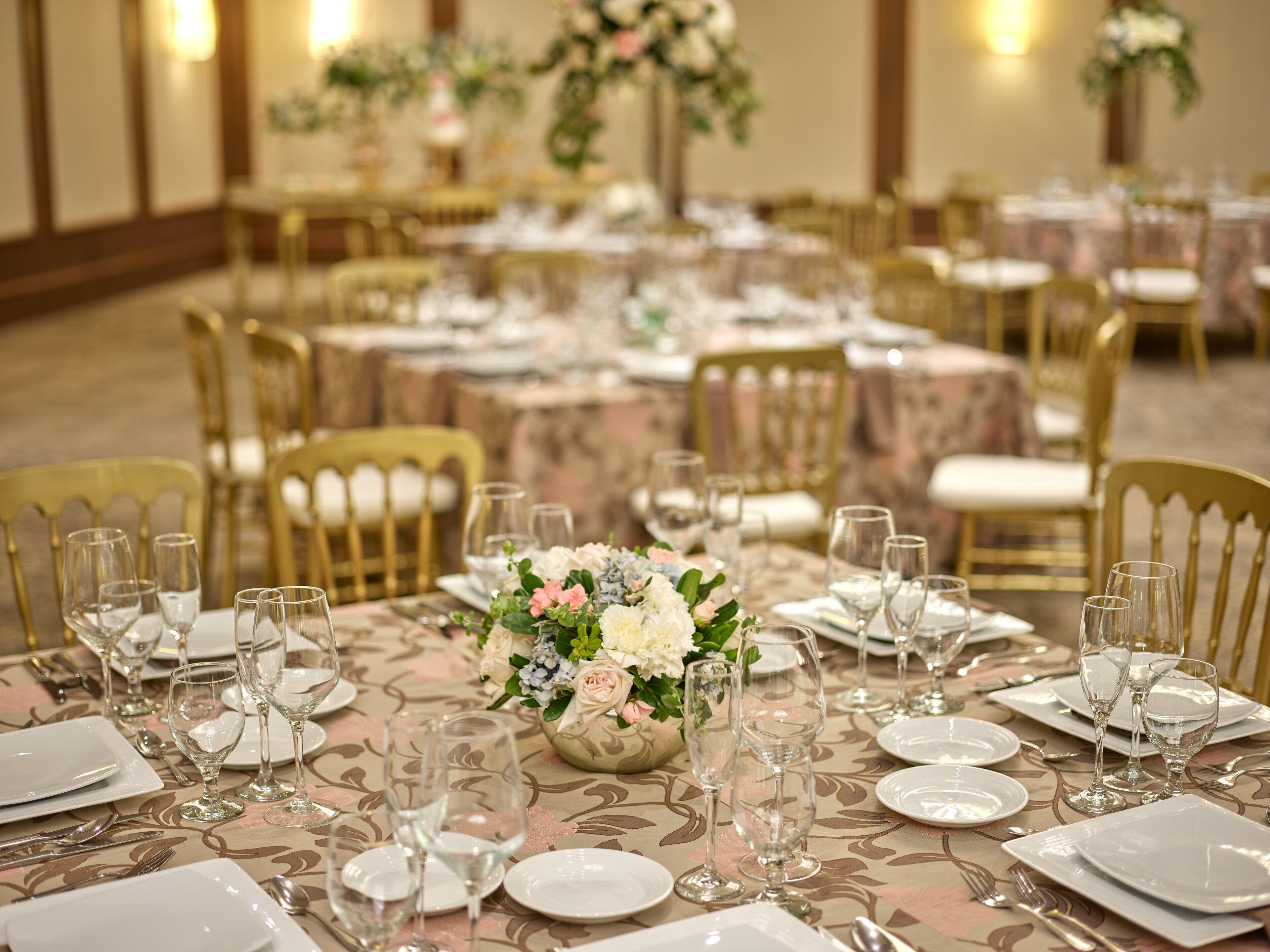 A banquet hall with round tables set for a formal event, featuring floral centerpieces and gold chairs.