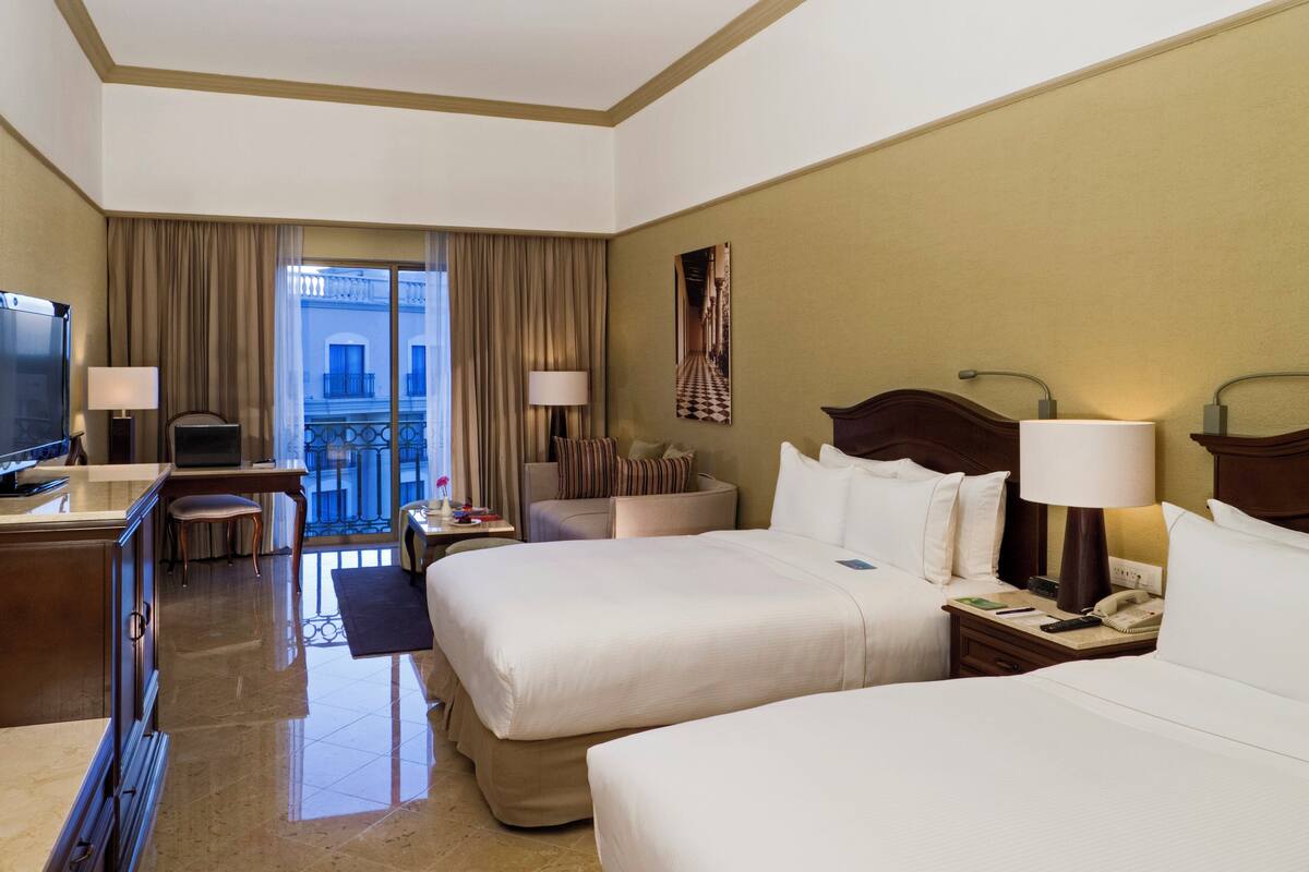 A hotel room with two beds, a desk with a television, a chair, a small table, and a view of a balcony.