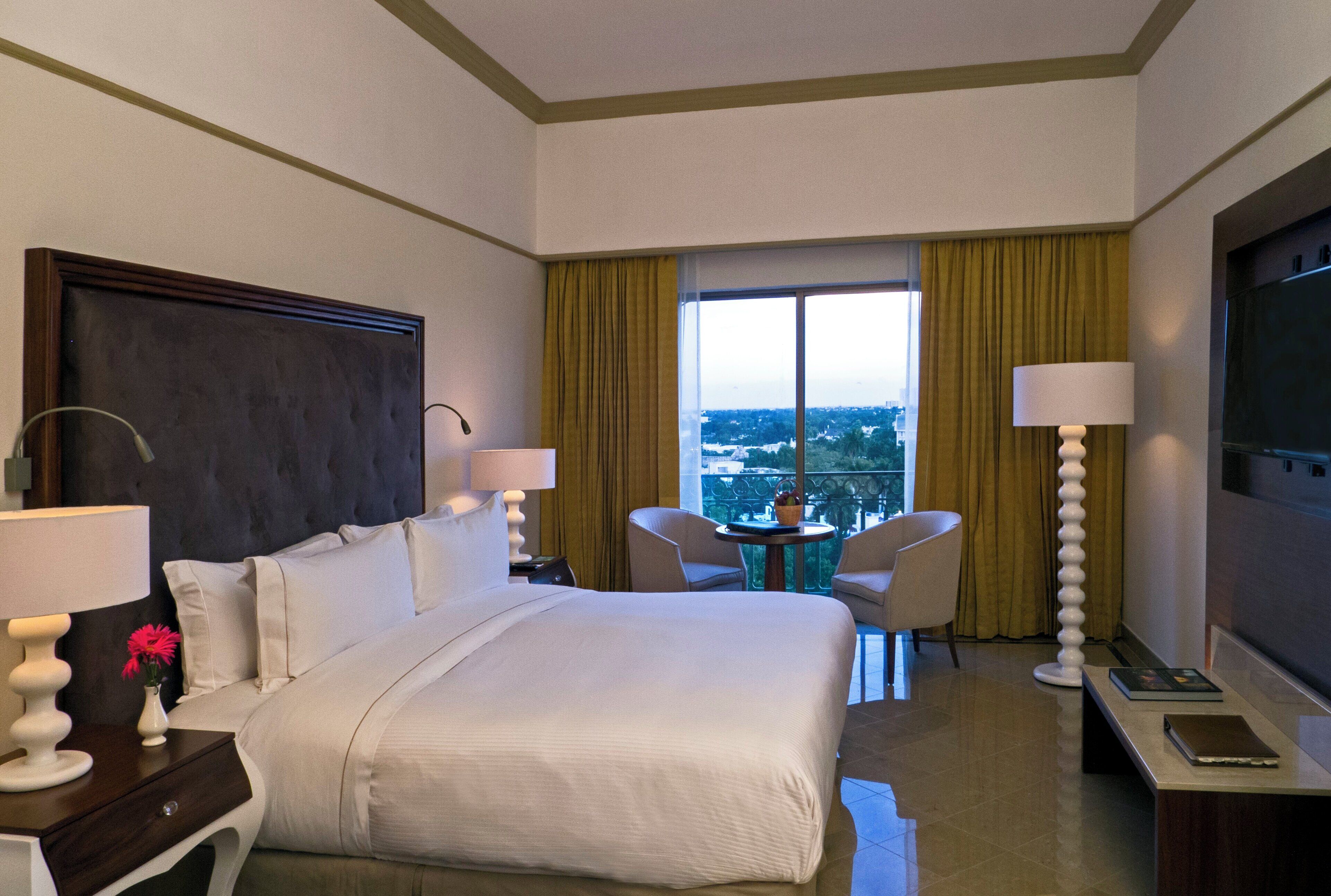 A hotel room with a large bed, two chairs, a small table, a TV, and a window with a city view.
