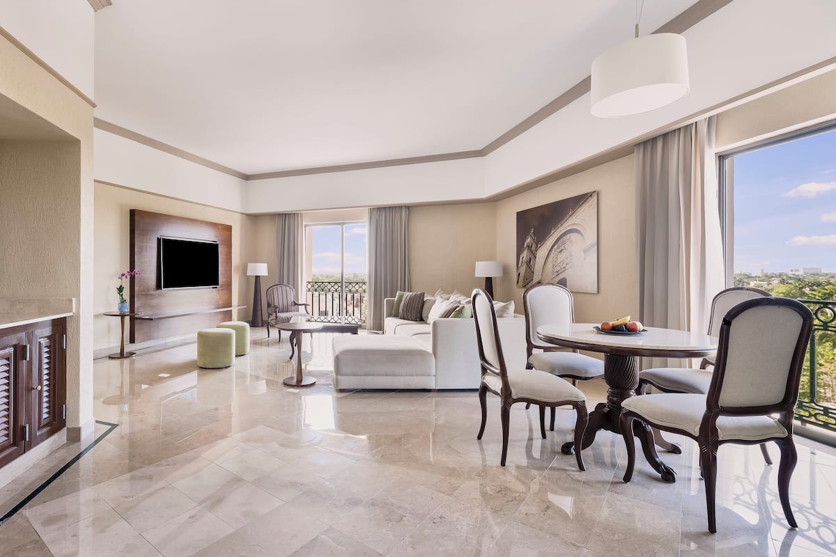 A modern hotel room with a living area, dining table, and a balcony with a city view.
