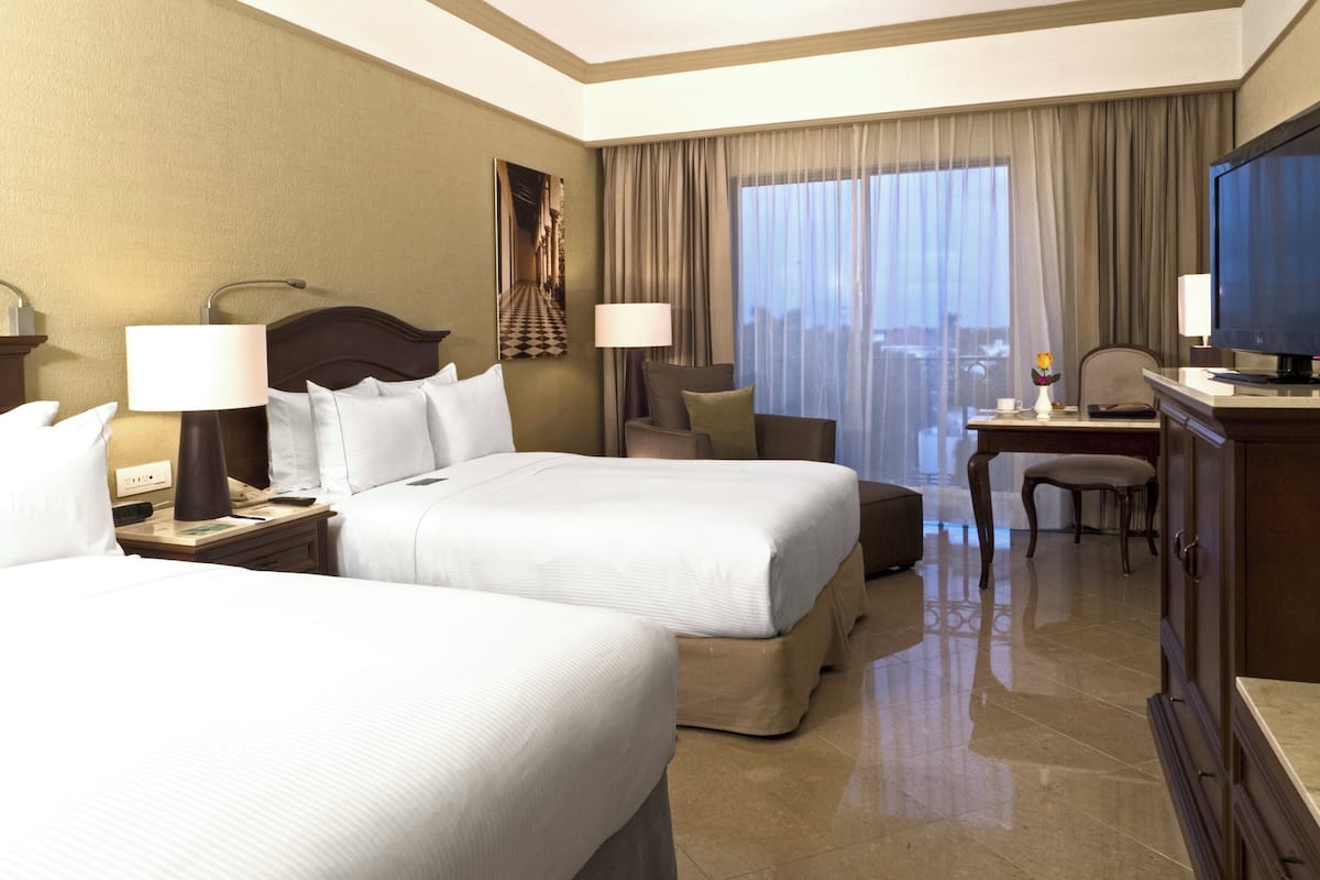 A hotel room with two beds, a desk, a TV, and a view of the city through large windows.