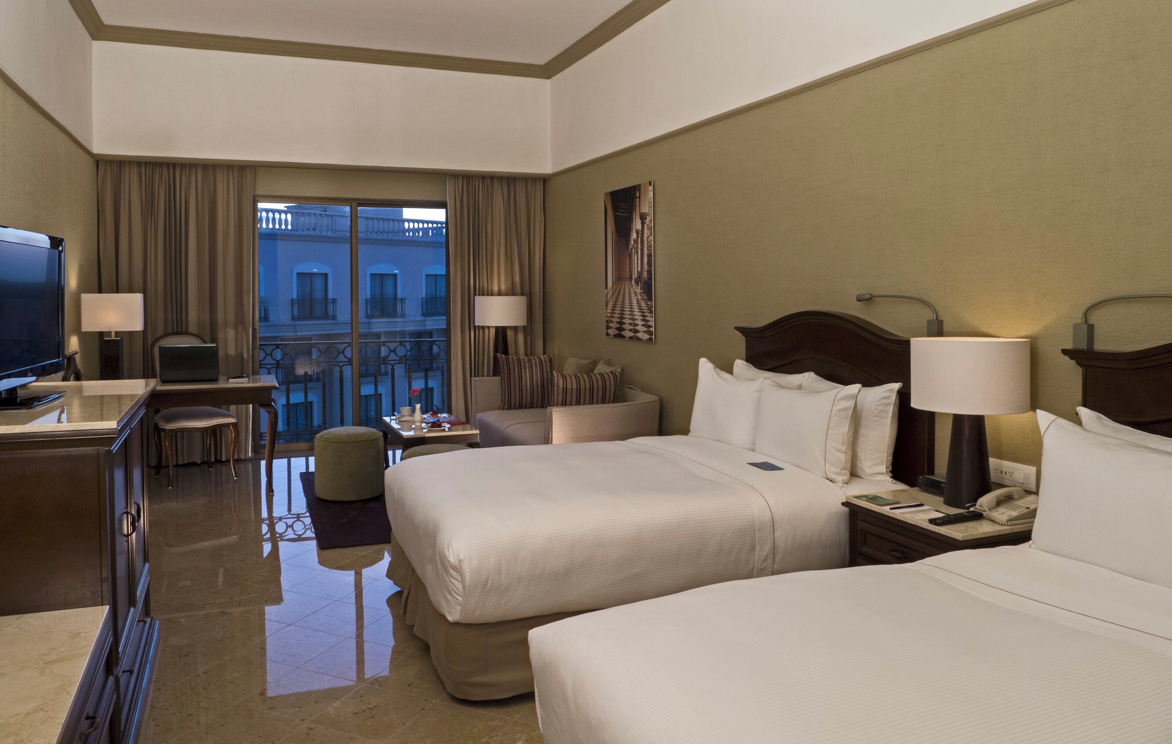 A hotel room with two beds, a desk with a TV, a chair, a small table, and a balcony with a view of buildings.