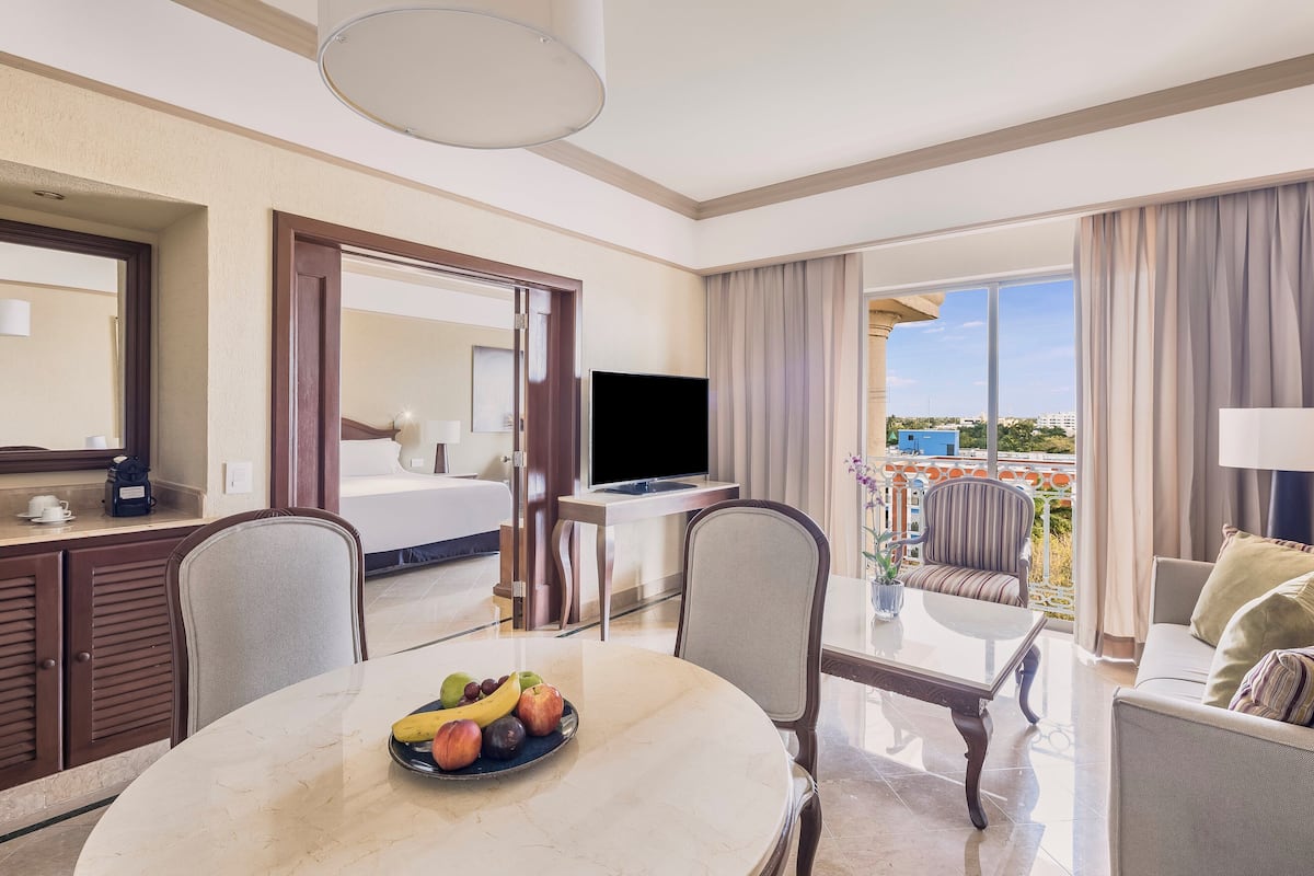A hotel room with a dining area, a TV, a balcony with a pool view, and a bed.