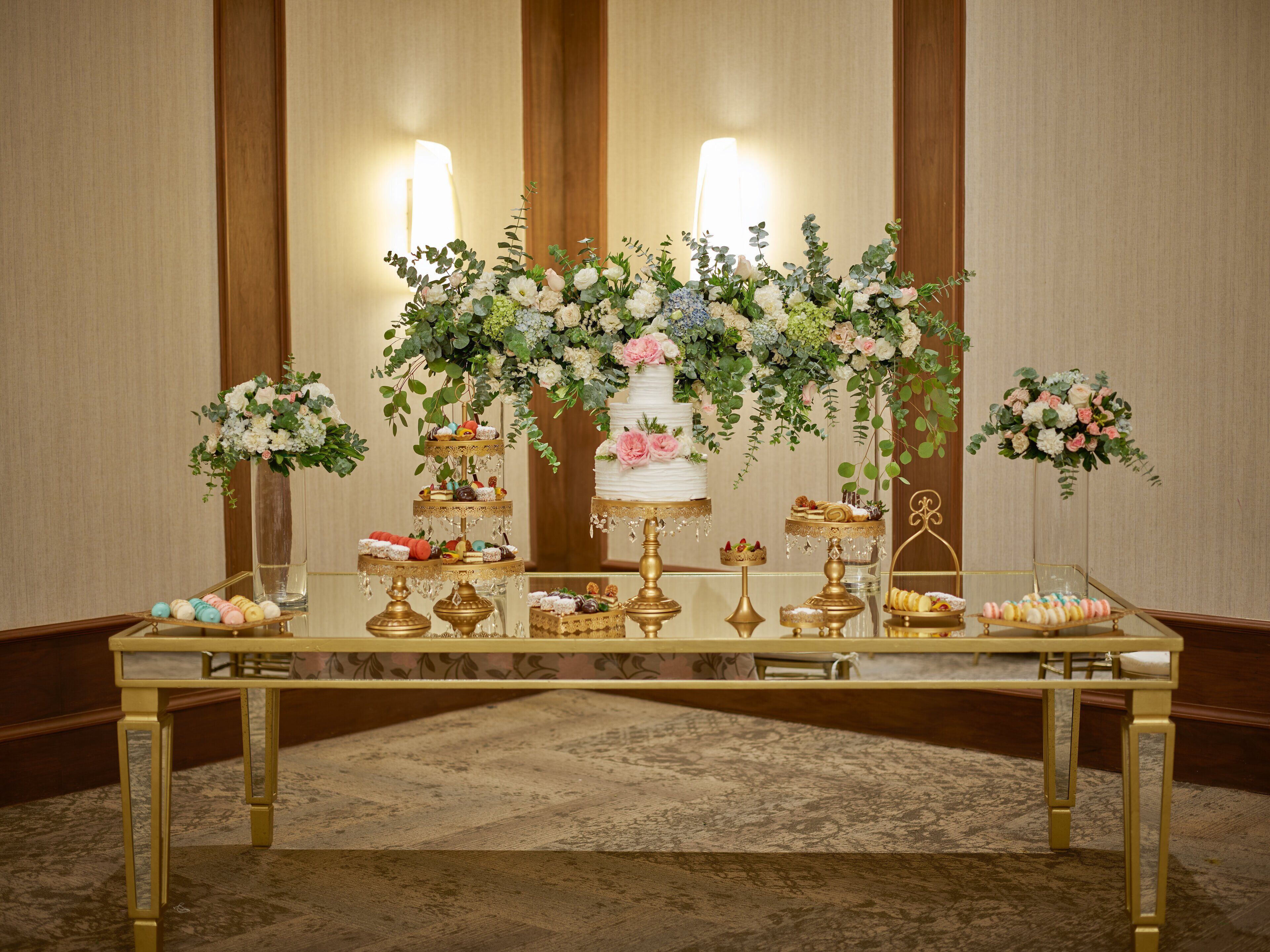 Una mesa de banquete con fuentes de postres escalonadas, arreglos florales y una torta nupcial de tres pisos.