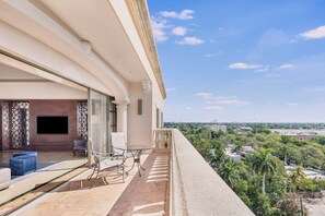 A balcony with a glass partition, a seating area with a table and chairs, and a view of a cityscape with greenery.