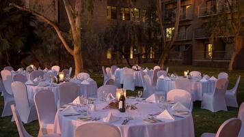 An outdoor banquet setup with round tables, white tablecloths, and chairs, surrounded by trees and a building in the background.