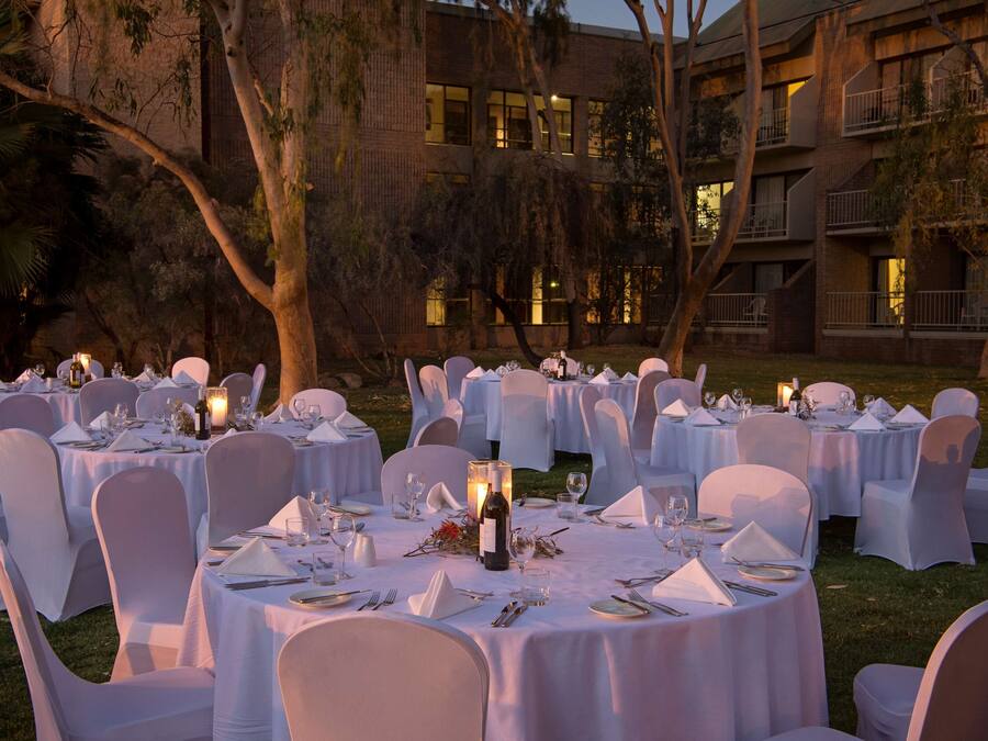An outdoor banquet setup with round tables, white tablecloths, and chairs, surrounded by trees and a building in the background.