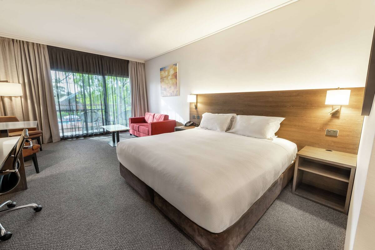 A hotel room with a large bed, a desk with a chair, a red armchair, and a window with curtains.