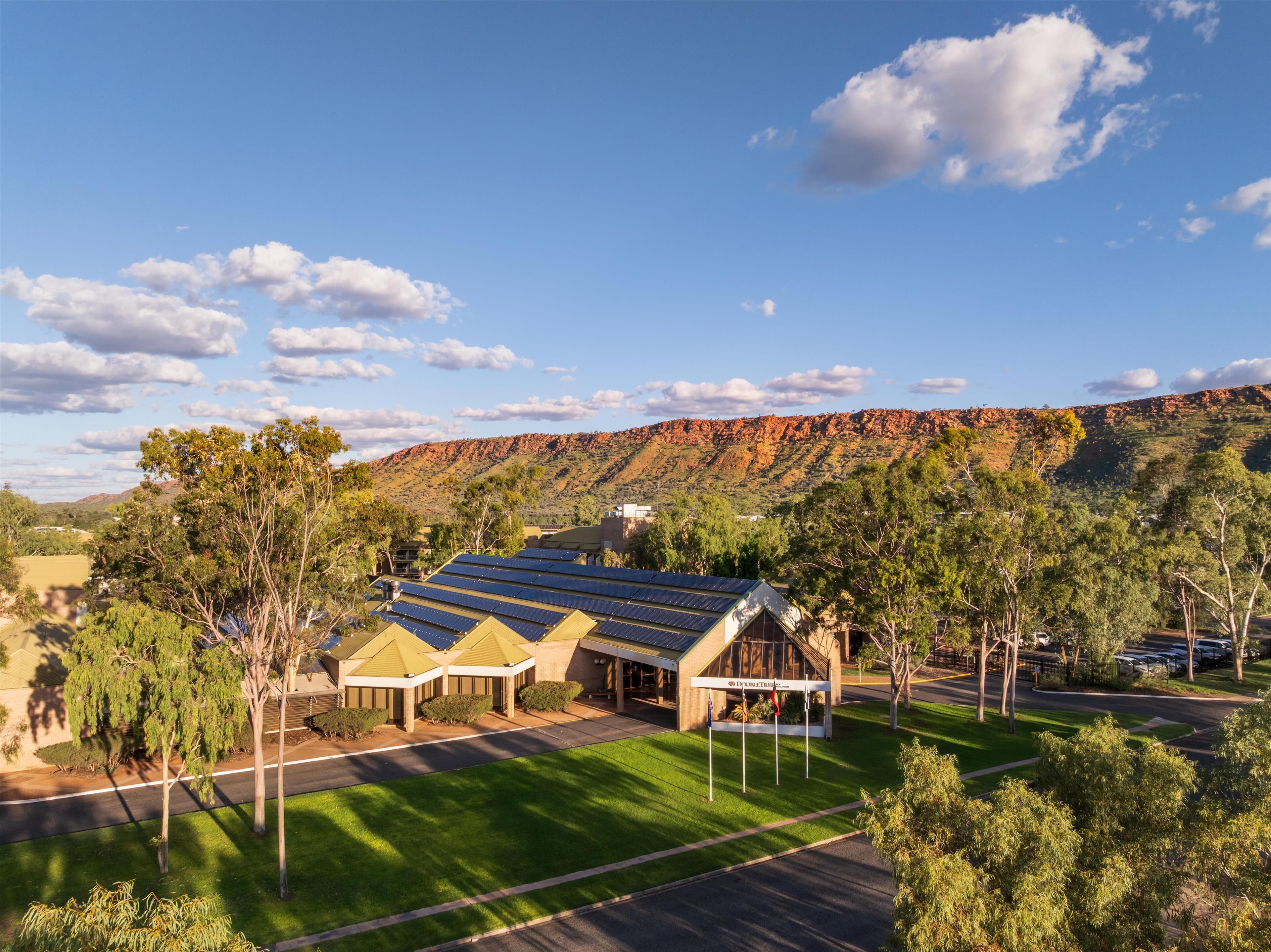 A building with solar panels, surrounded by trees and a hilly landscape.