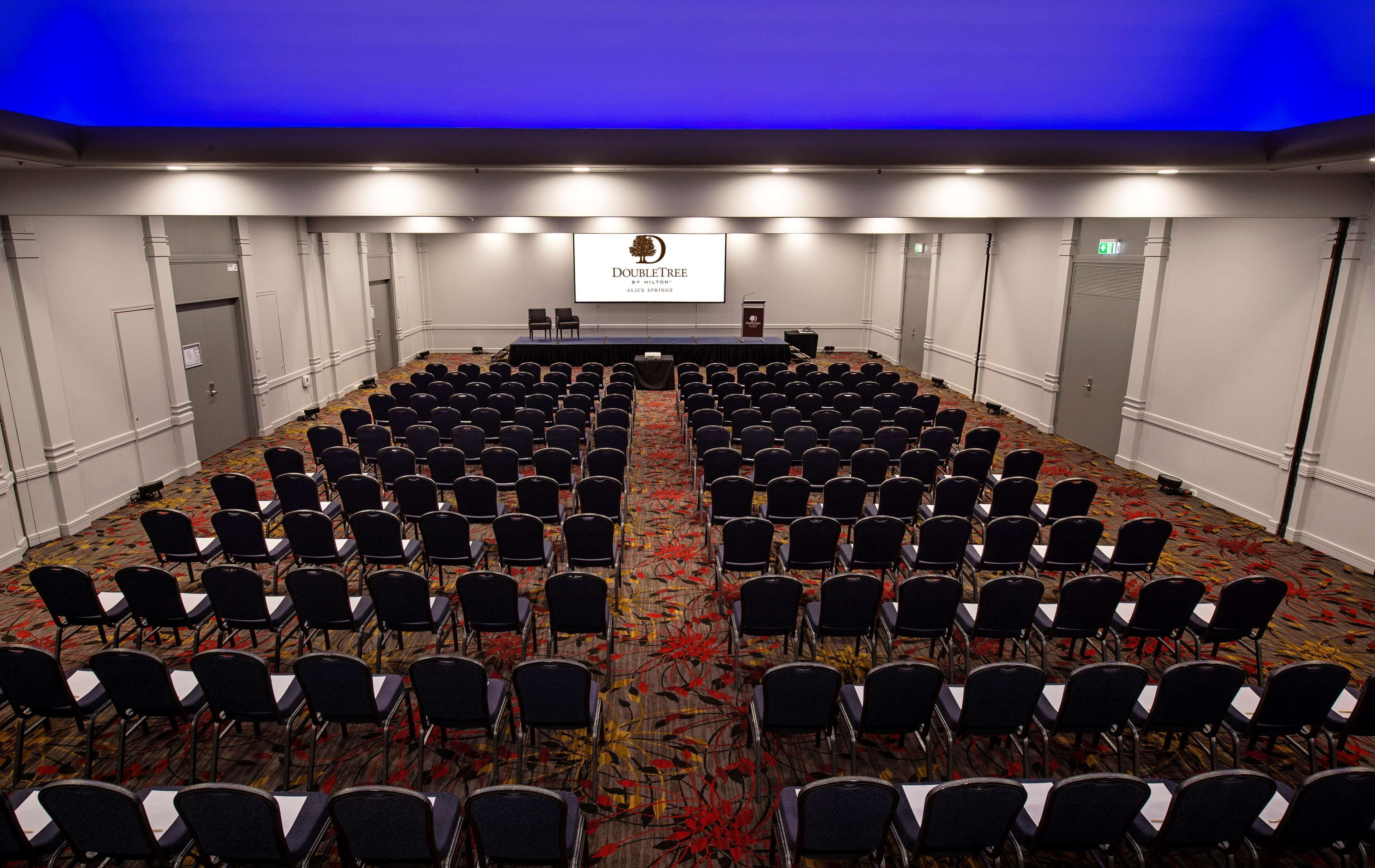 Un salón de conferencias con filas de sillas, un atril y una pantalla que muestra 
