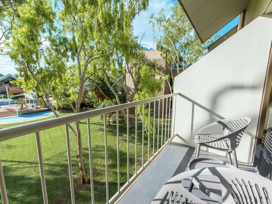 A balcony with a metal railing, a pool, and a view of a building and trees.