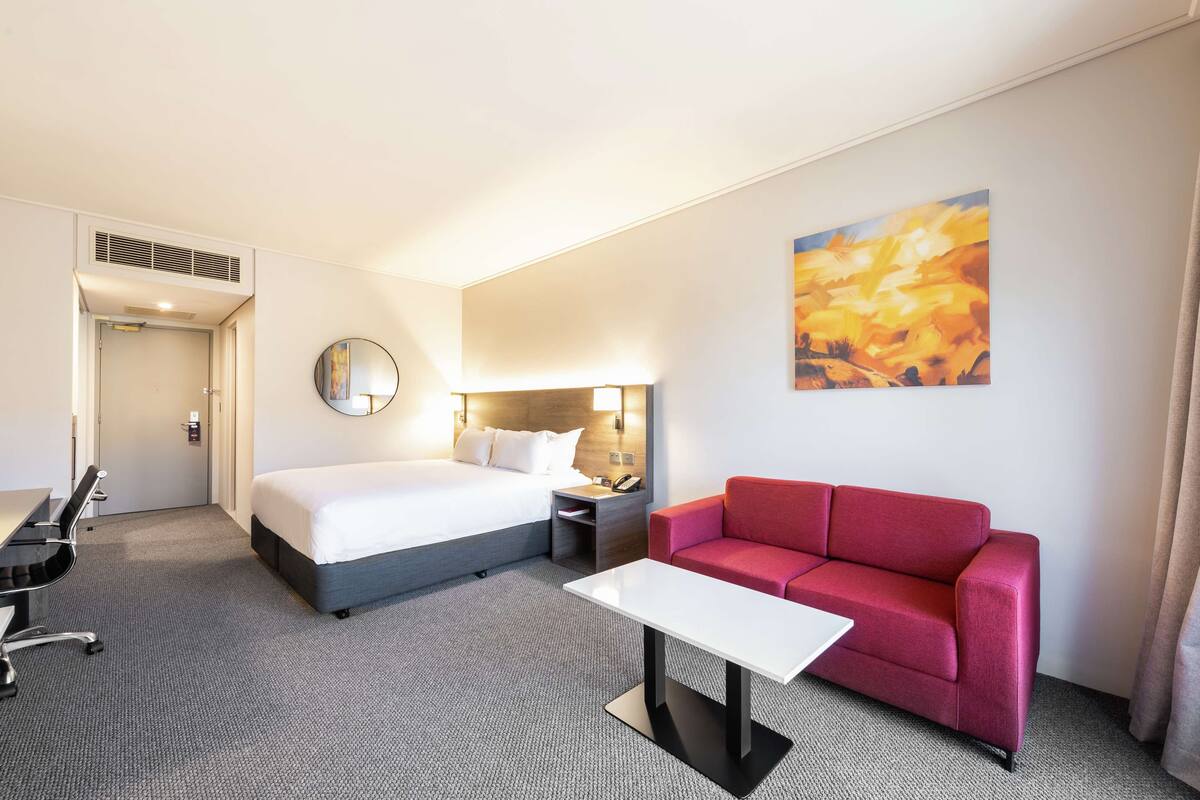 A hotel room with a bed, a red sofa, a white table, a desk with a chair, a round mirror, and a painting on the wall.