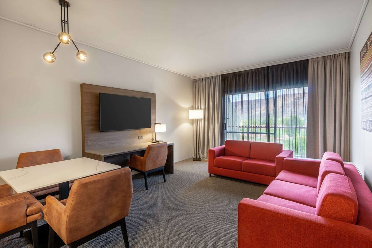 A hotel room with a dining table, chairs, a red sofa, a TV, and a large window with curtains.