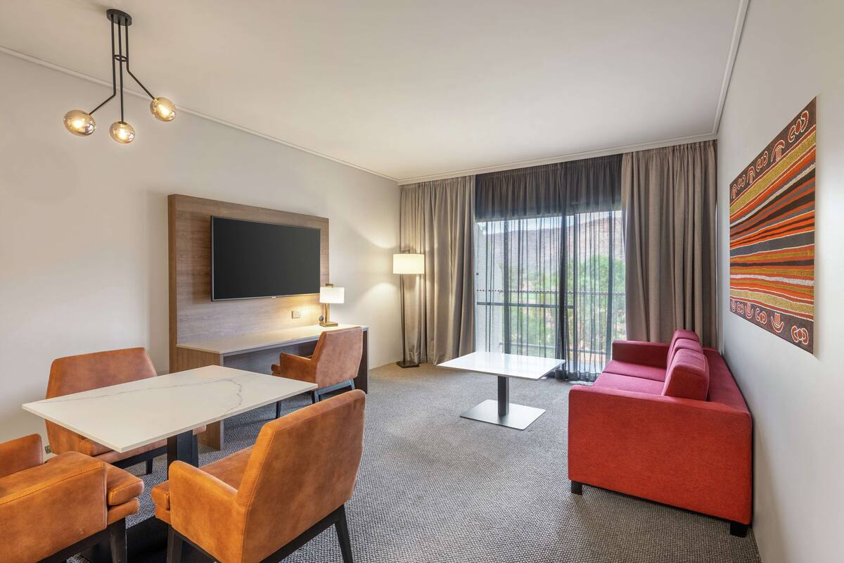 A hotel room with a dining table, chairs, a sofa, a TV, and a large window with curtains.