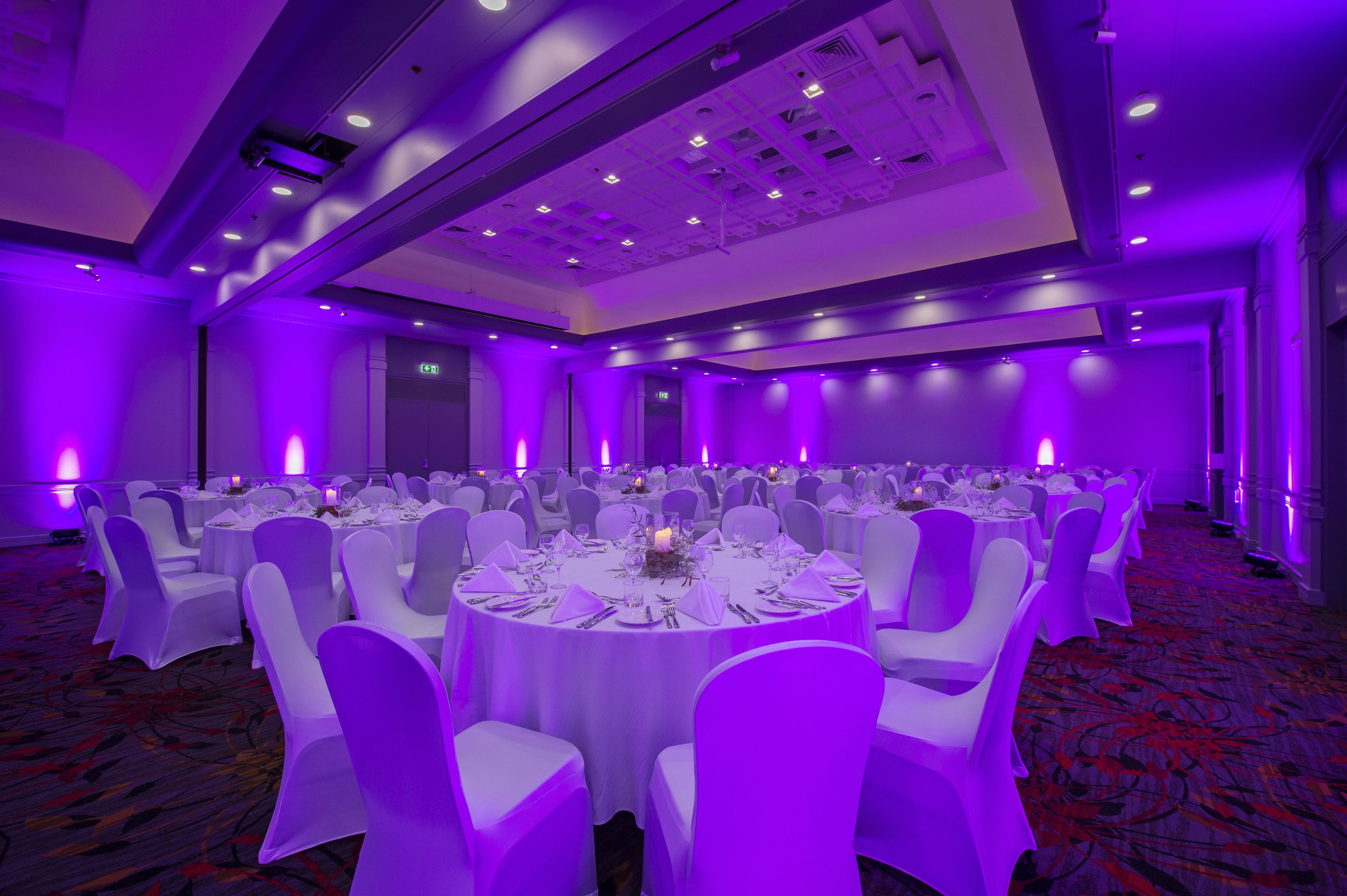 A banquet hall with round tables set for a formal event, featuring white tablecloths and chairs, and purple ambient lighting.