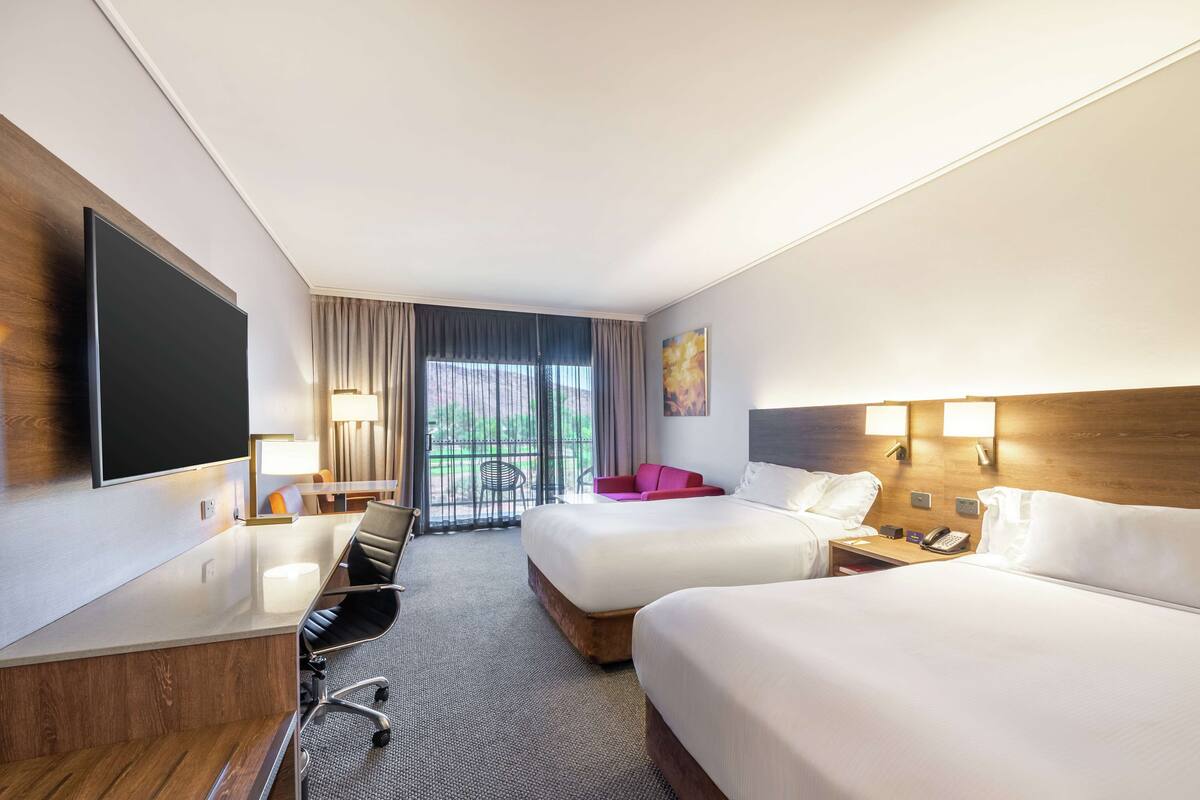 A hotel room with two beds, a desk, a flat-screen TV, a chair, a lamp, and a window with curtains.
