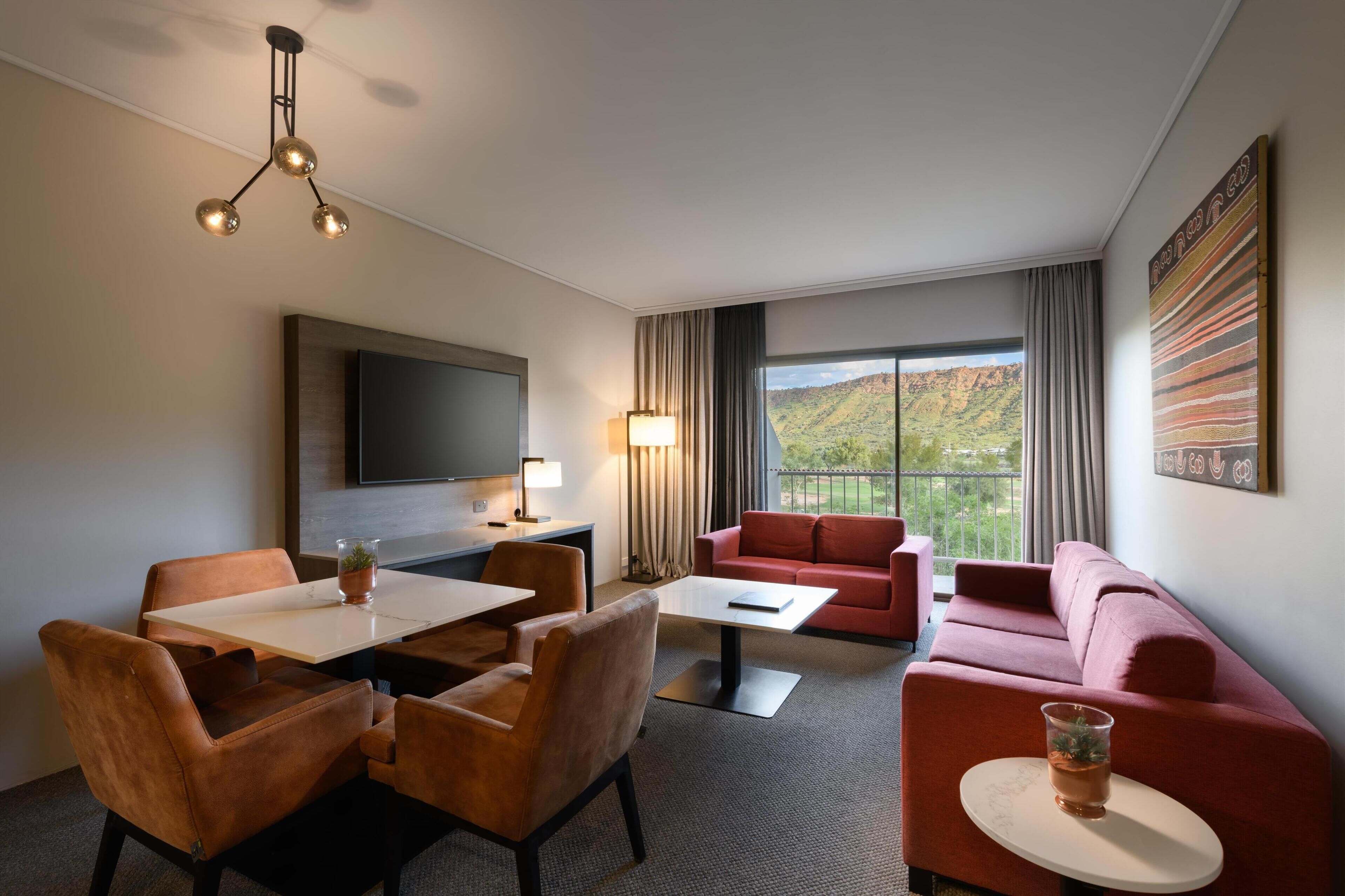 A hotel room with a dining area, a sofa, a TV, and a large window with a mountain view.