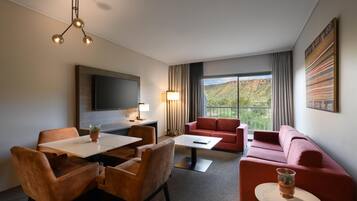 A hotel room with a dining area, a sofa, a TV, and a large window with a mountain view.