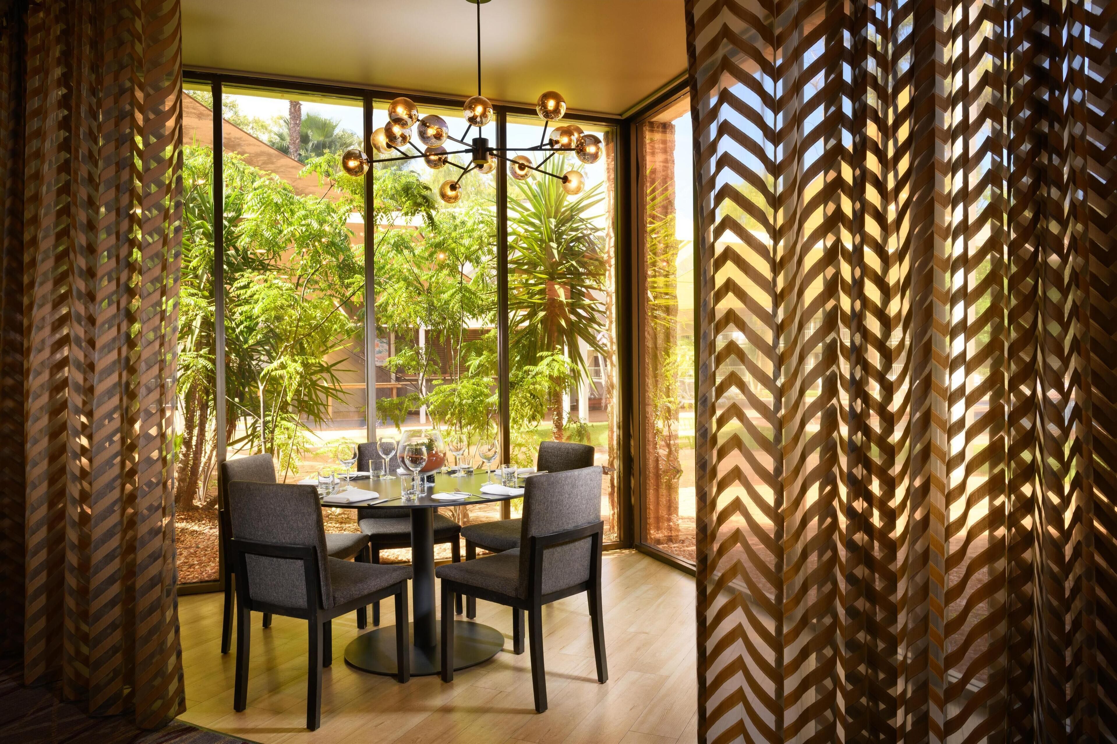 A dining area with a table set for four, surrounded by chairs, with a view of greenery outside.