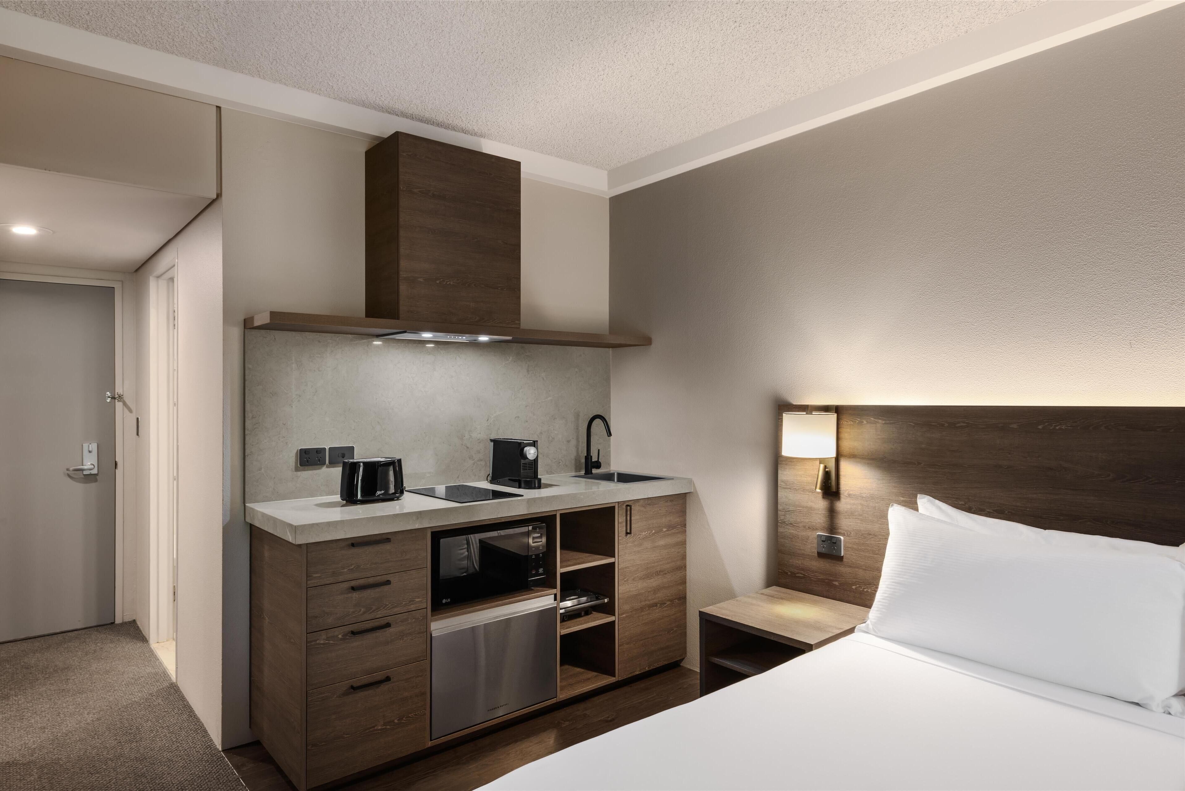 A compact hotel room with a kitchenette, a bed with white linens, a nightstand with a lamp, and a wooden headboard.