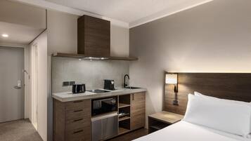 A compact hotel room with a kitchenette, a bed with white linens, a nightstand with a lamp, and a wooden headboard.