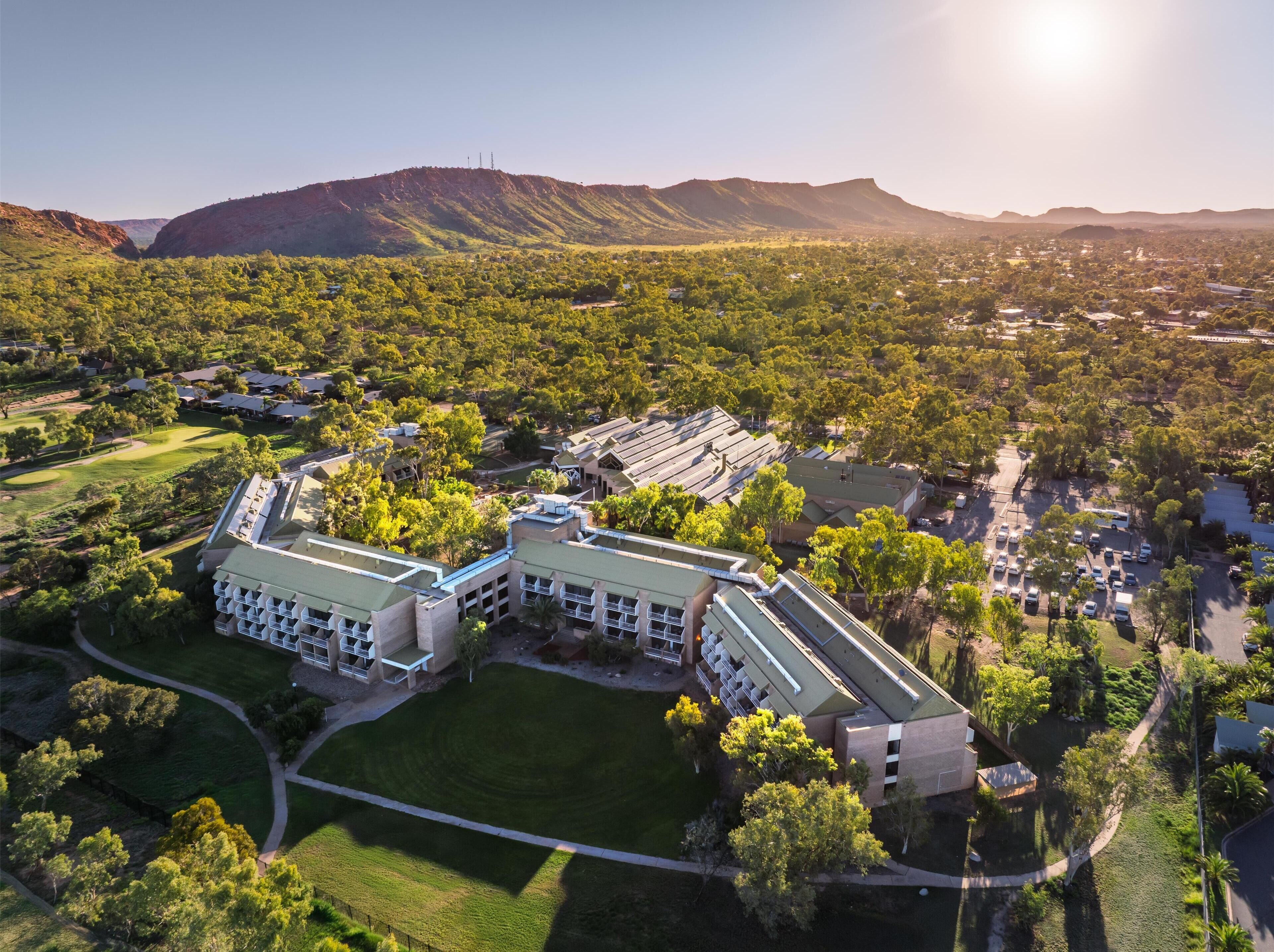 An aerial view of a large, modern hotel with multiple buildings, surrounded by lush greenery and mountains in the background.