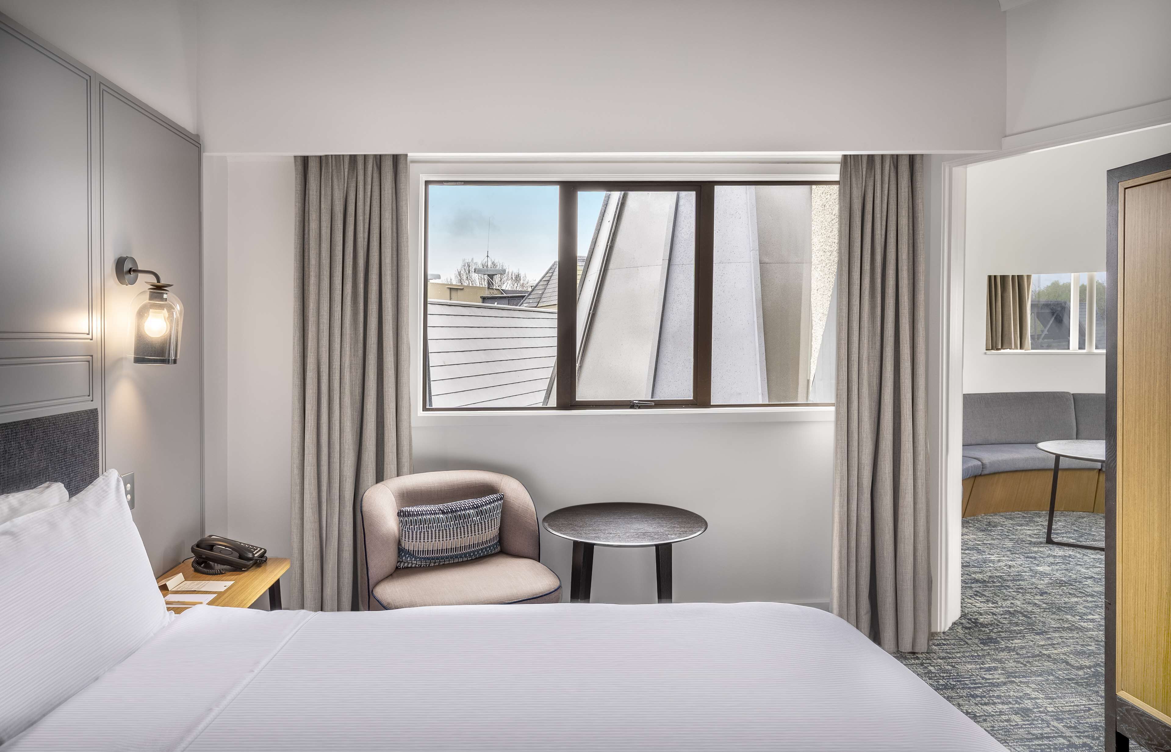 Een hotelkamer met een groot bed, een klein rond tafeltje, een stoel, een nachtkastje, een raam met gordijnen en uitzicht op het omliggende landschap.