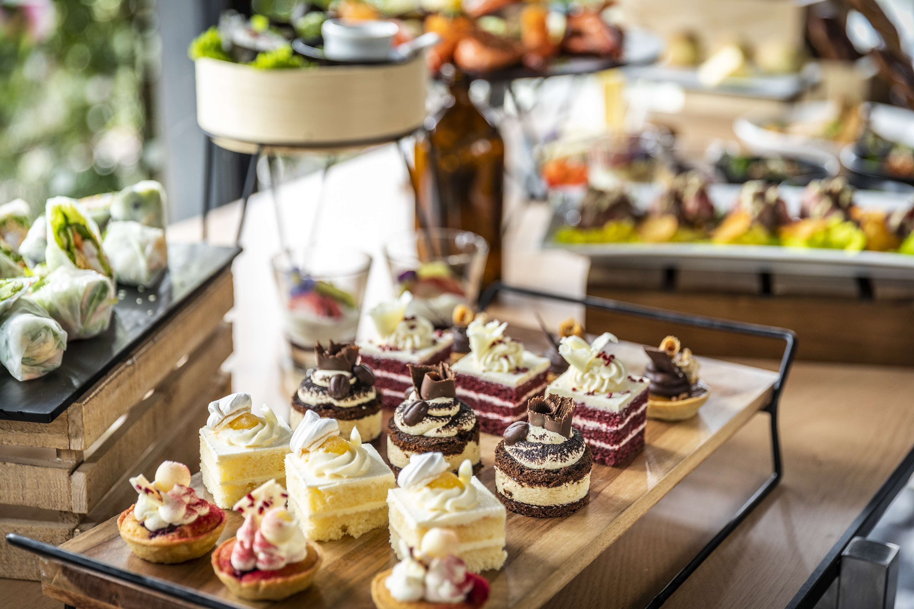 A variety of desserts on a wooden board, including cakes and pastries, with a blurred background of additional food items.
