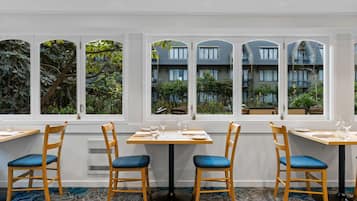 Un restaurante con vistas a un edificio a través de amplias ventanas, mesas de madera y sillas con cojines azules.